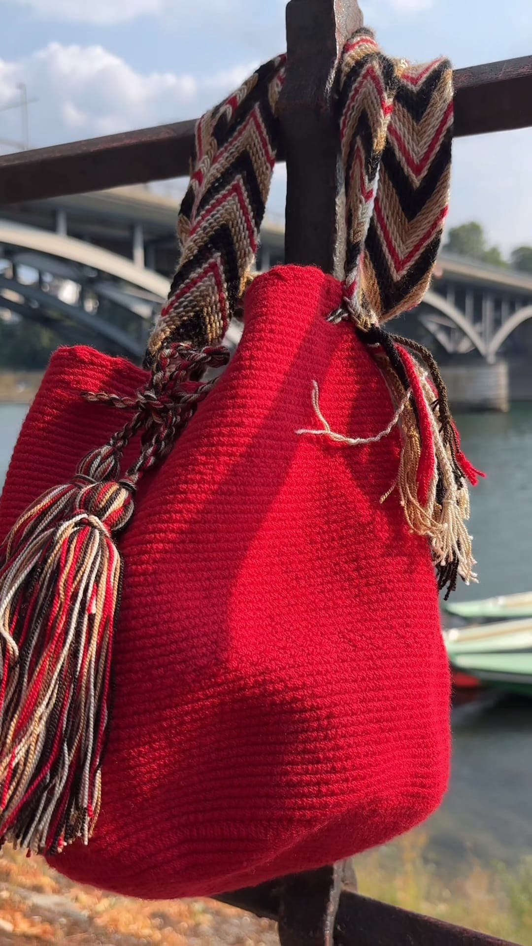 Handgewebte rote Wayuu-Mochila mit gemustertem Gurt hängt an einem Geländer am Rhein in Basel, im Hintergrund die Wettsteinbrücke